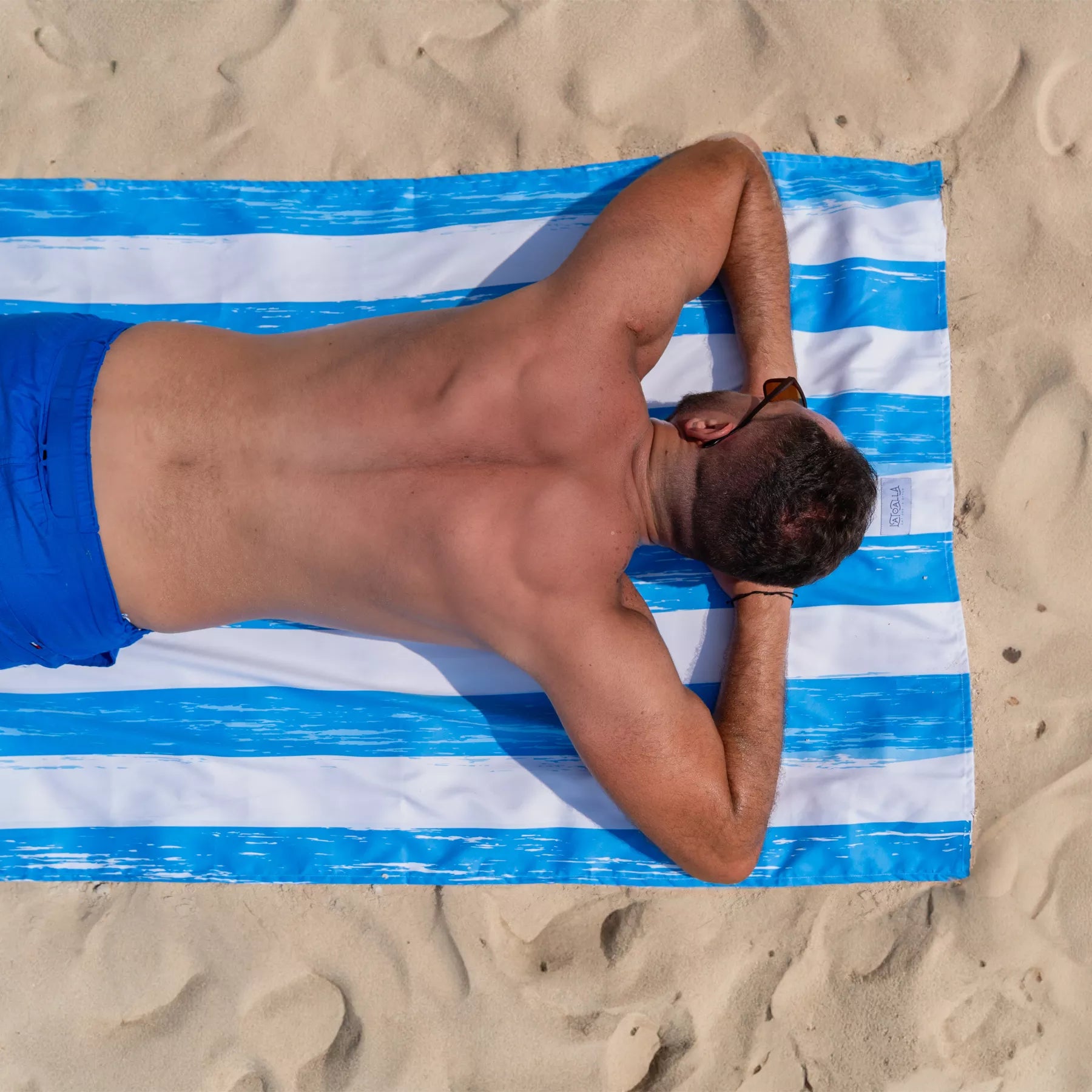 A guy sitting on a Quick Dry, Sand Free, Light Weight, Compact and Ecofriendly Microfiber Beach Towel from La Toalla on the sand