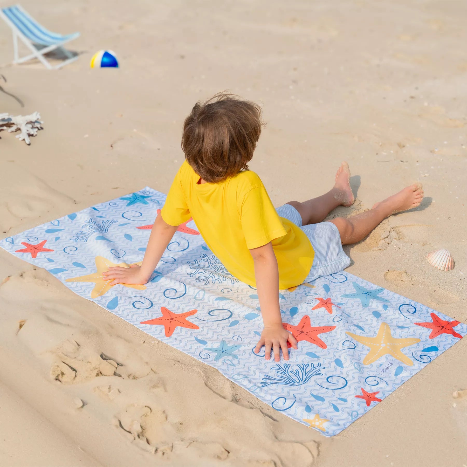 A boy sitting on the sand on a Quick Dry, Sand Free, Light Weight, Compact and Ecofriendly Microfiber Beach Towel from La Toalla designed for kids