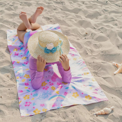 A girl sitting on the beach on a Quick Dry, Sand Free, Light Weight, Compact and Ecofriendly Microfiber Beach Towel from La Toalla with kids design